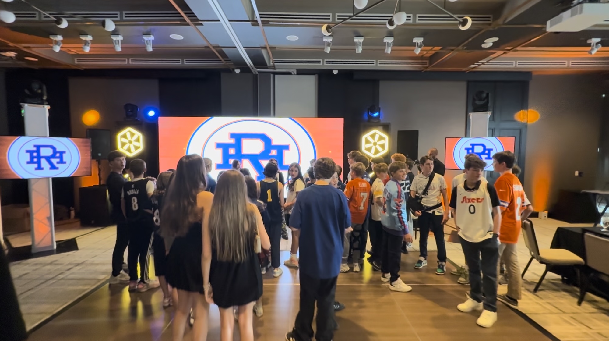 Basketball-Themed Bar Mitzvah Gathering Group of young people at a basketball-themed event with sports jerseys and a large screen displaying basketball-related graphics.