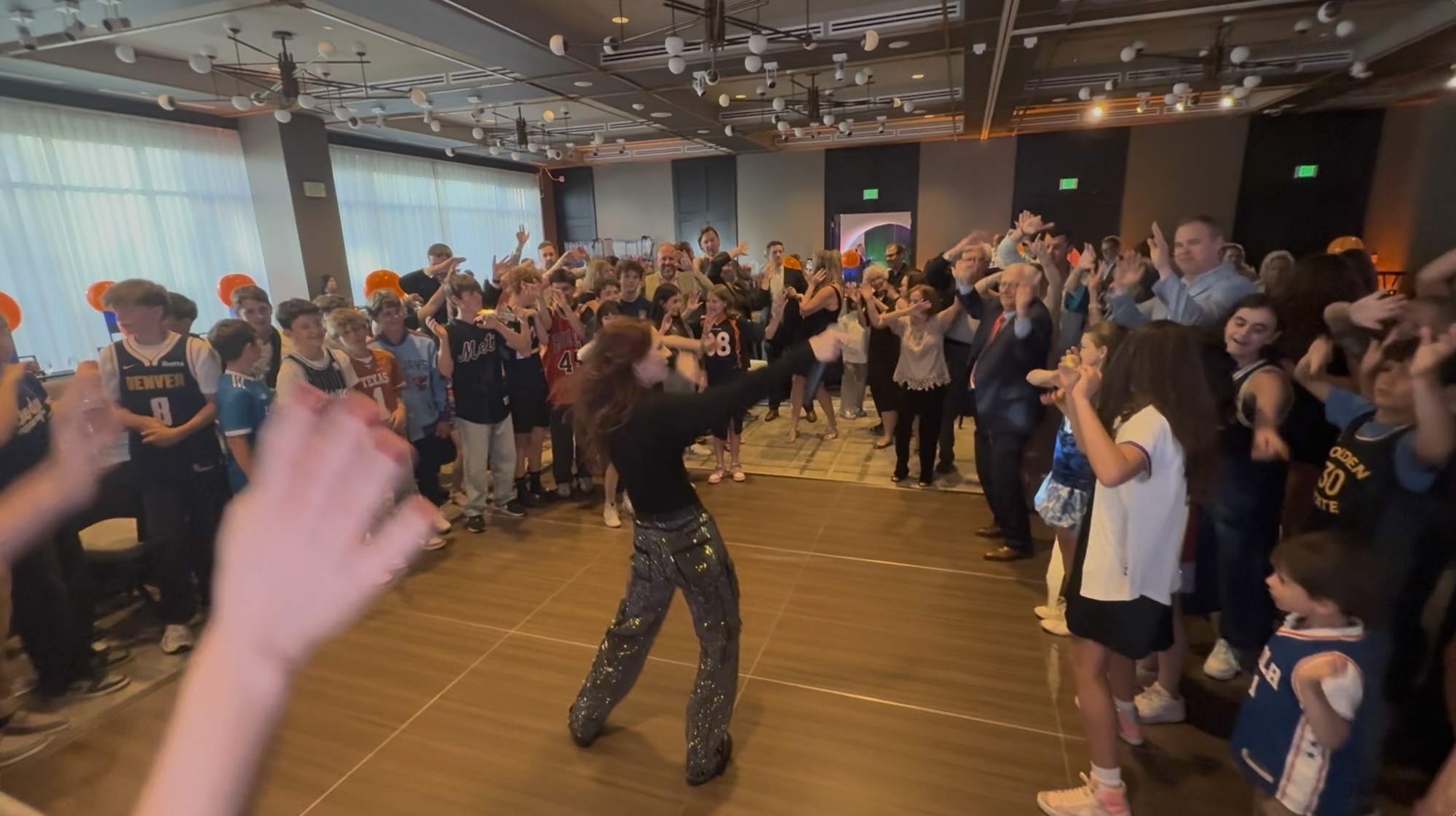 Screenshot Group of people dancing and celebrating at a basketball-themed event in a spacious, well-lit venue.
