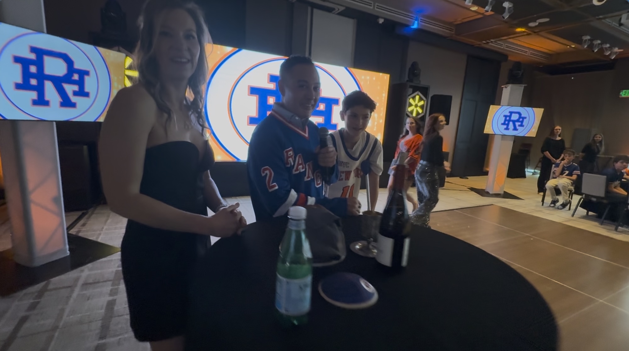 Hank's Basketball Themed Bar Mitzvah Event People gathered around a table at an event with large video screens in the background.