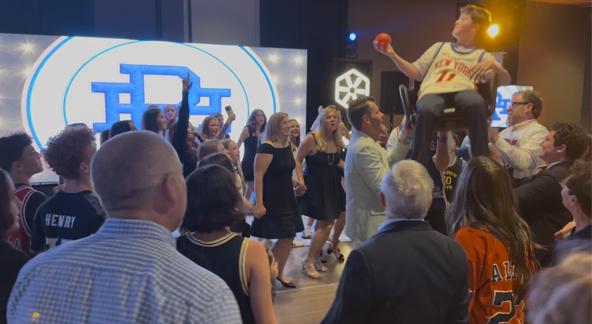 Basketball Themed Bar Mitzvah Celebration A crowd of people dancing and celebrating with a young boy hoisted on a chair, holding a basketball, during a party.