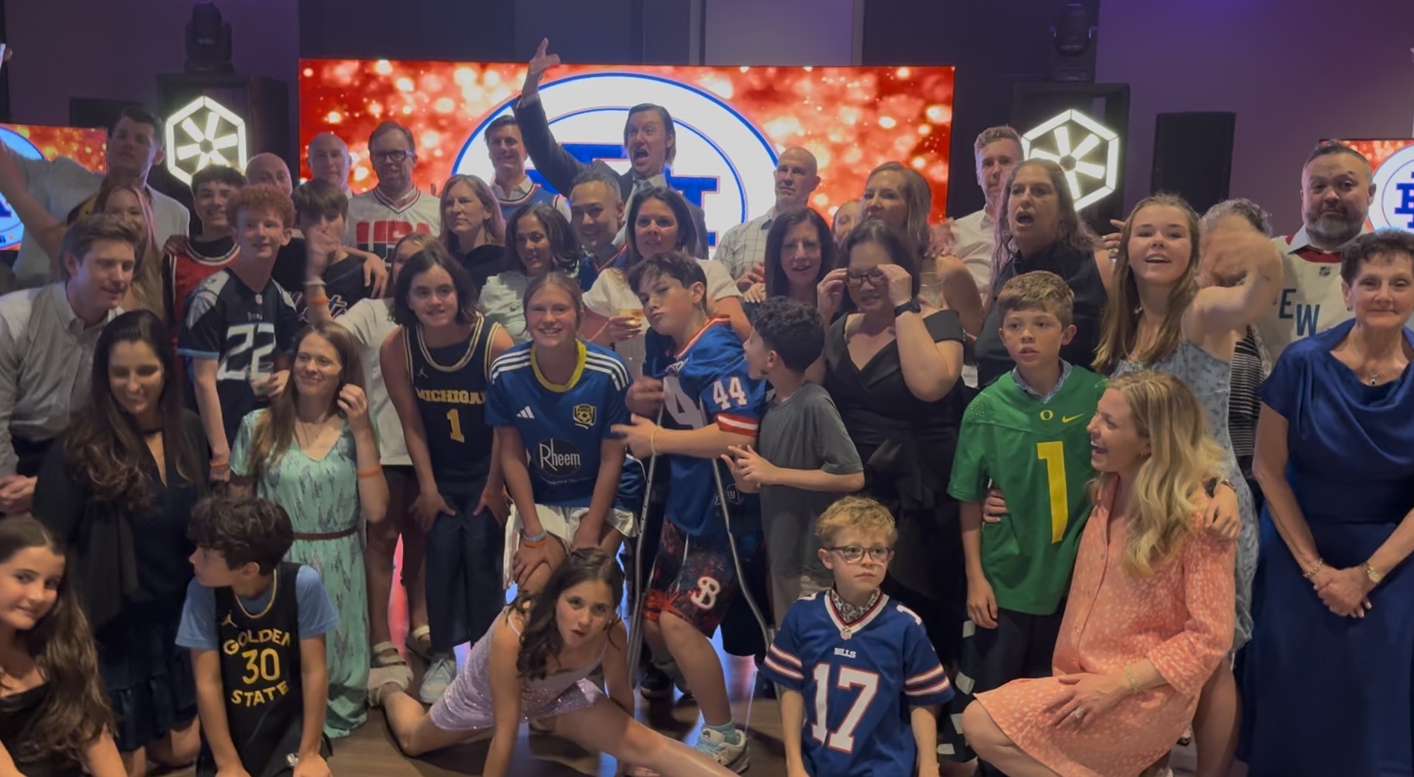 Hank's Basketball Themed Bar Mitzvah Group of people at a celebration, with some wearing sports jerseys, posing in front of a colorful backdrop.