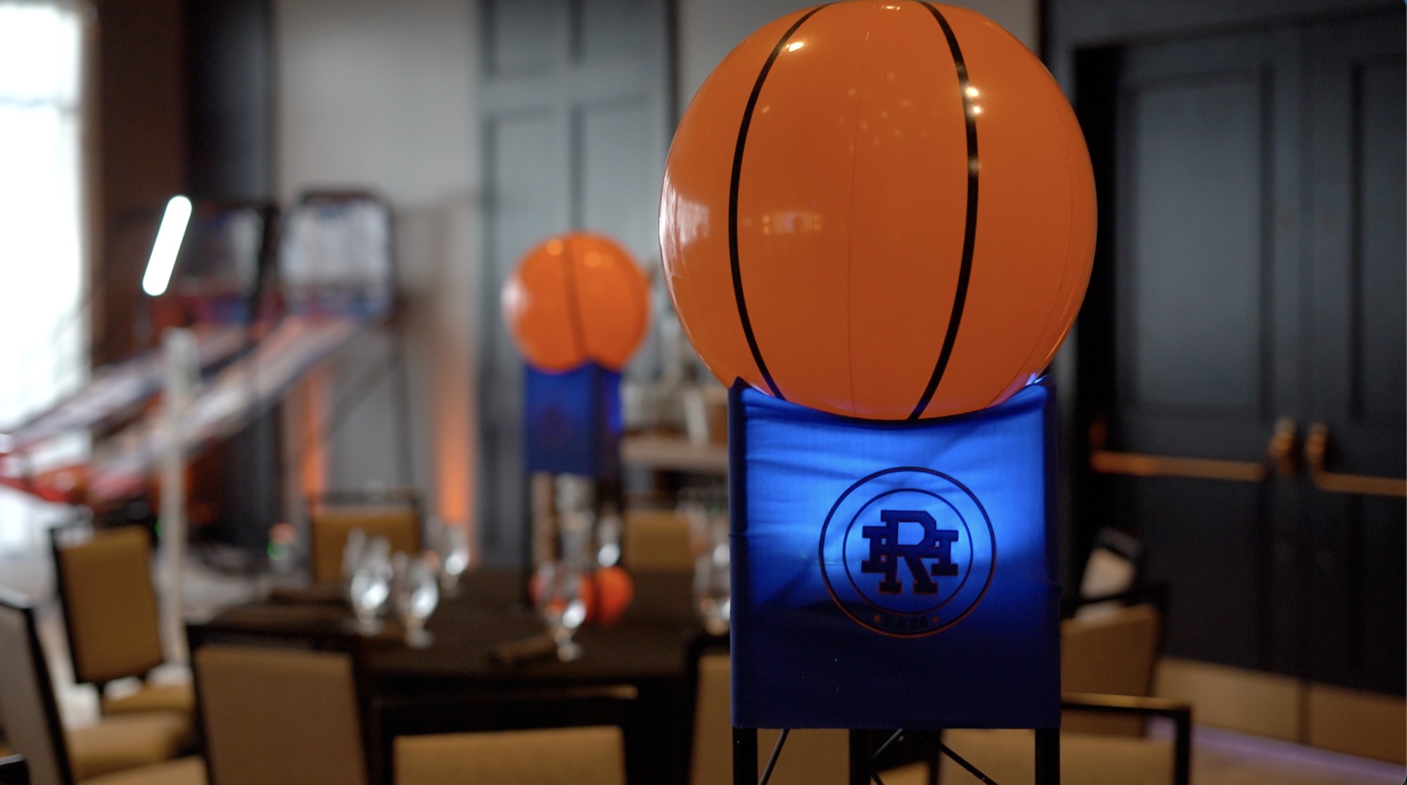 Screenshot An orange basketball decoration sits atop a blue stand with a logo, part of a basketball-themed event setup. In the background, tables and chairs are visible, along with blurred arcade games and another similar decoration.