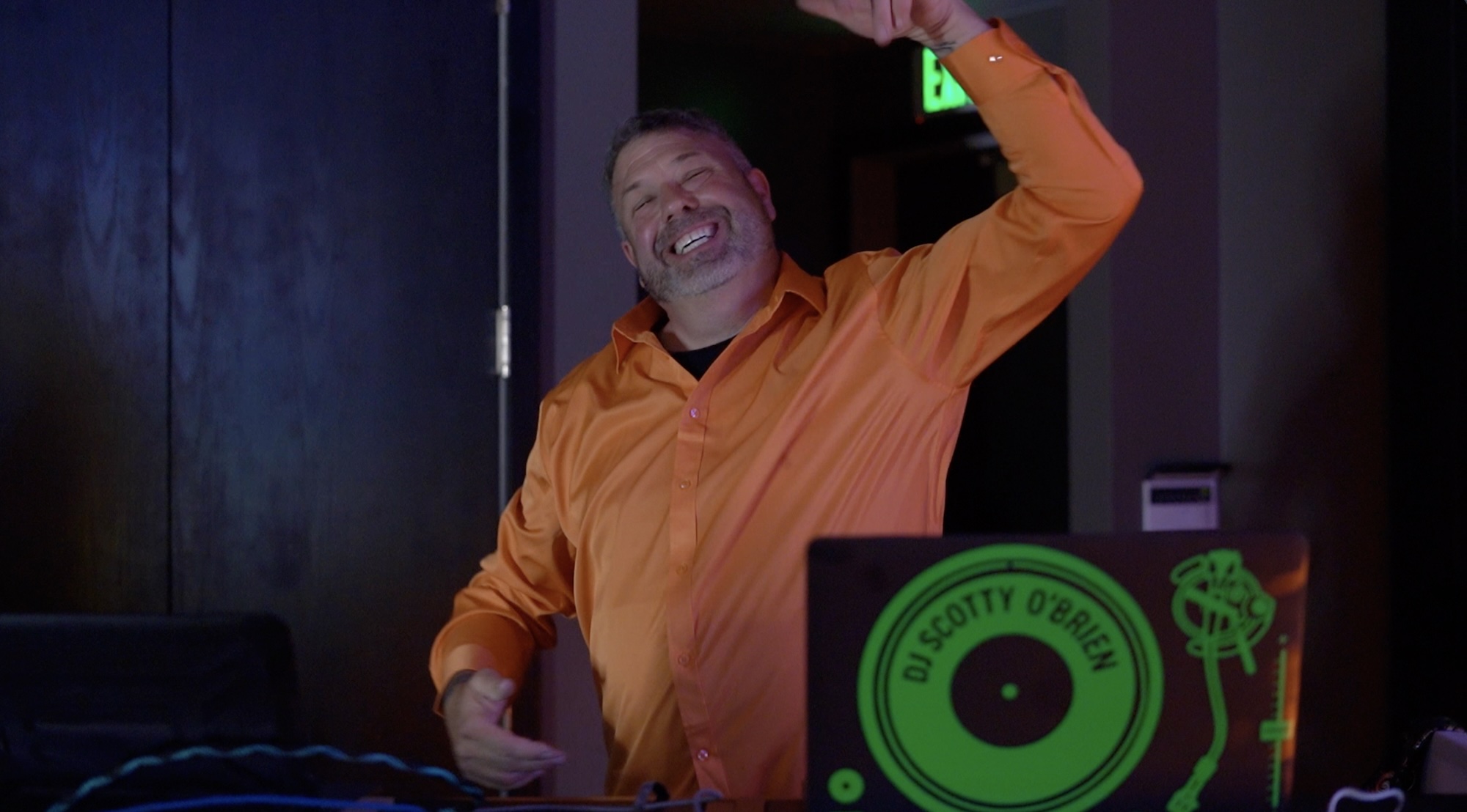DJ at Bar Mitzvah Celebration DJ in orange shirt smiling and gesturing energetically behind DJ booth at bar mitzvah.